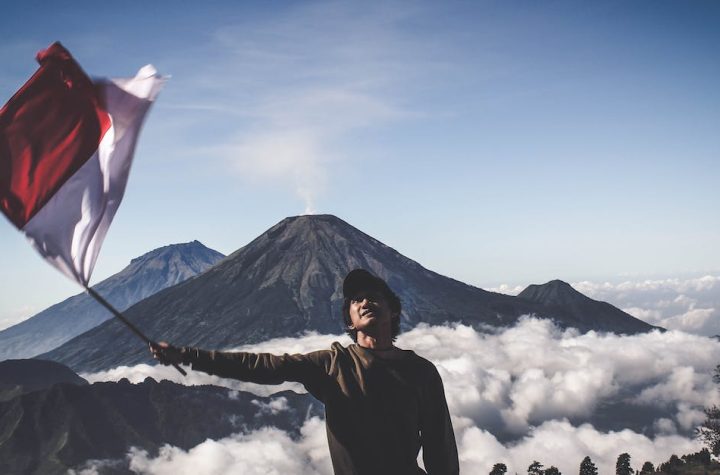 man-wearing-black-crew-neck-sweater-holding-white-and-red-flag-standing-near-mountain-under-blue-and